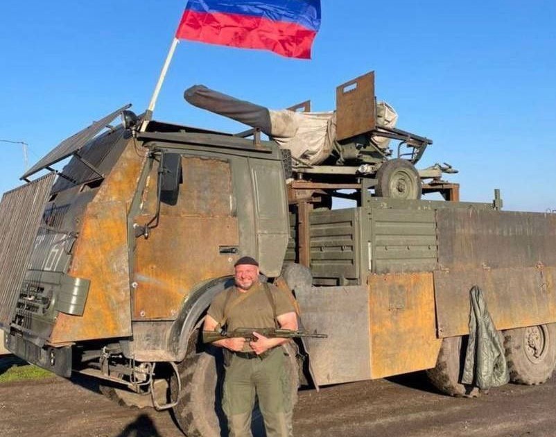 Una foto muestra a un corpulento invasor ruso sonriendo frente a otro cubo oxidado del ejército ruso que también ha sido muy modificado.